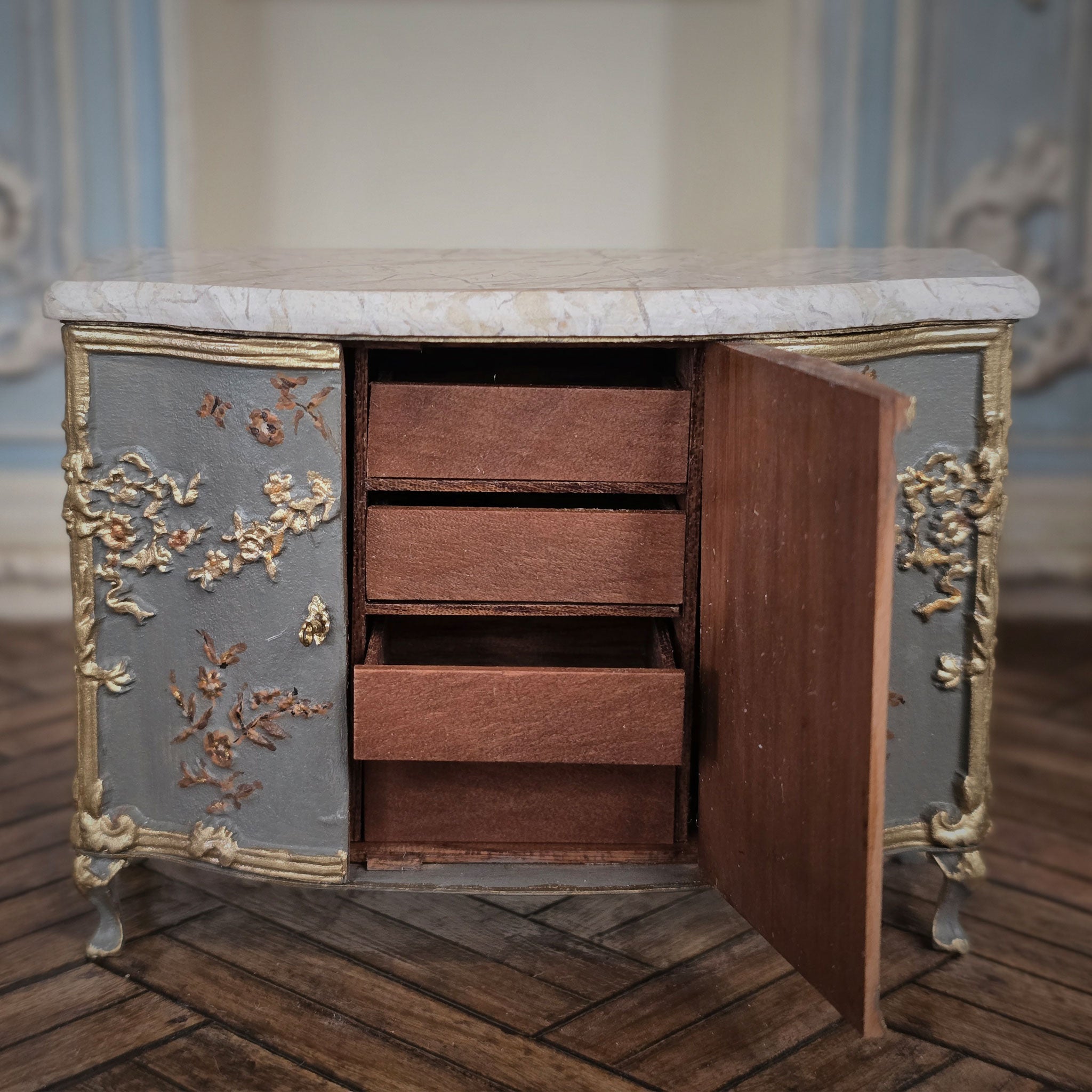 Interior of miniature sideboard cabinet with marble top and ornate gold detailing on a wooden floor.