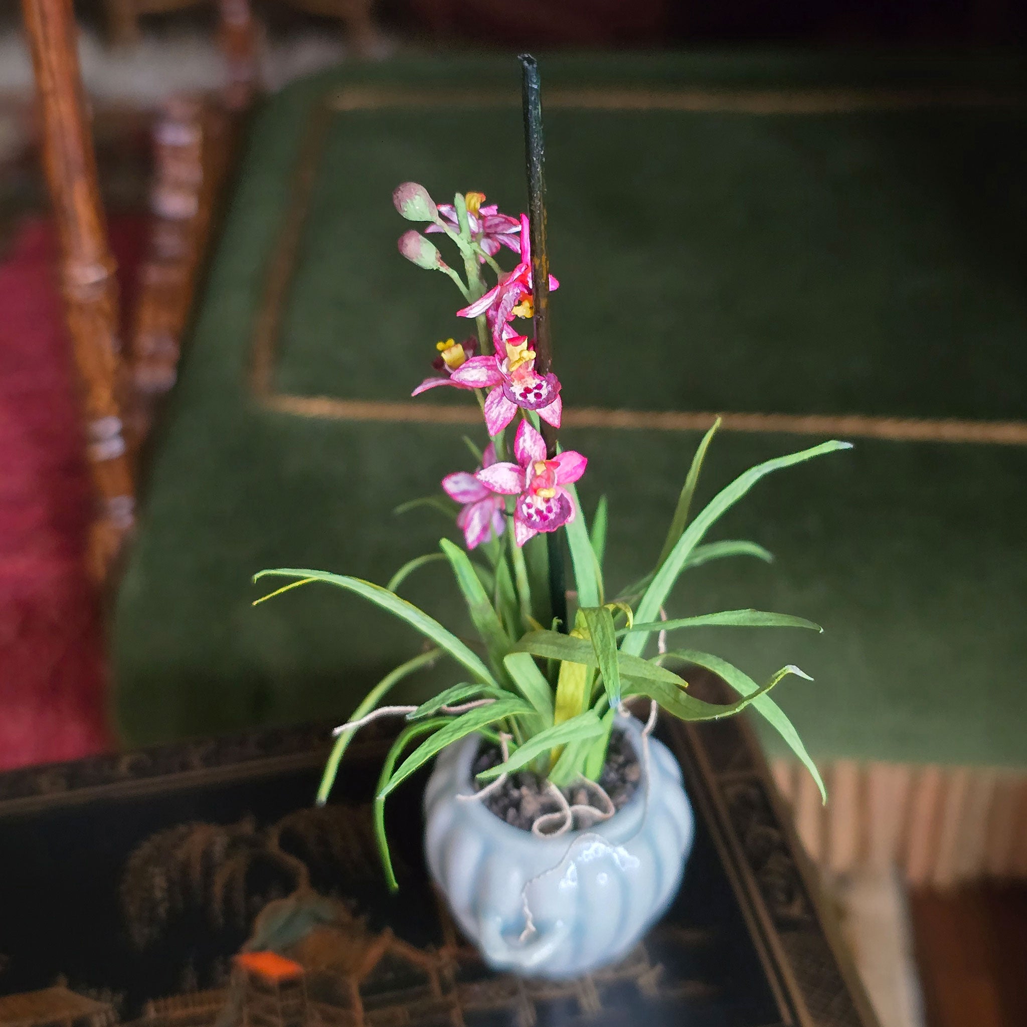 Potted orchid with hand painted paper pink petals and green leaves in a porcelain planter, on a table with a blurred background