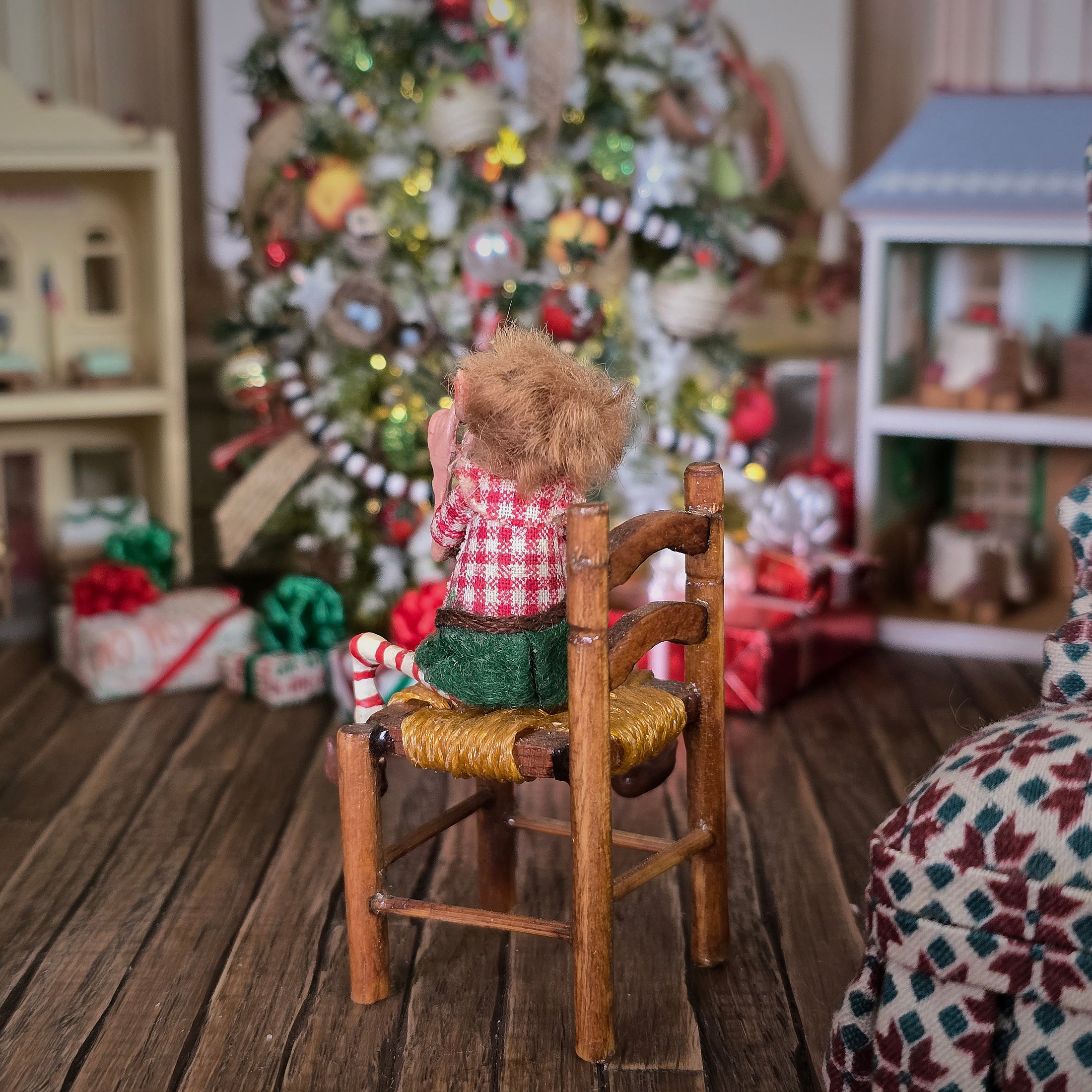 Grinning Elf Christmas Doll Cynthia Baron sitting on a wooden chair in a festive room with a Christmas tree and presents.