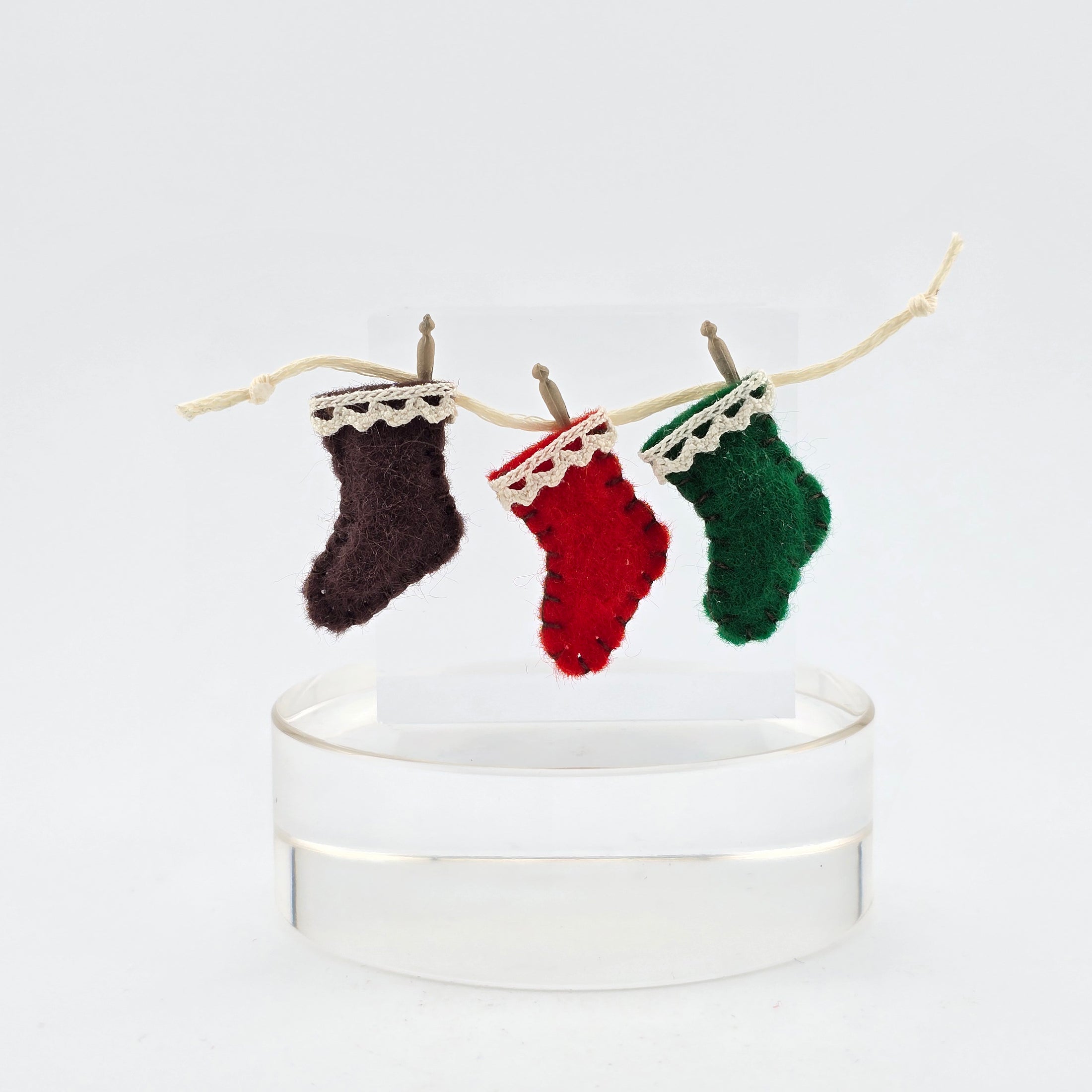 Three miniature wool stockings in brown, red, and green hanging on a string against a white background.