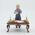 Load image into Gallery viewer, Miniature doll of a boy standing behind a table with various Christmas items on a white background
