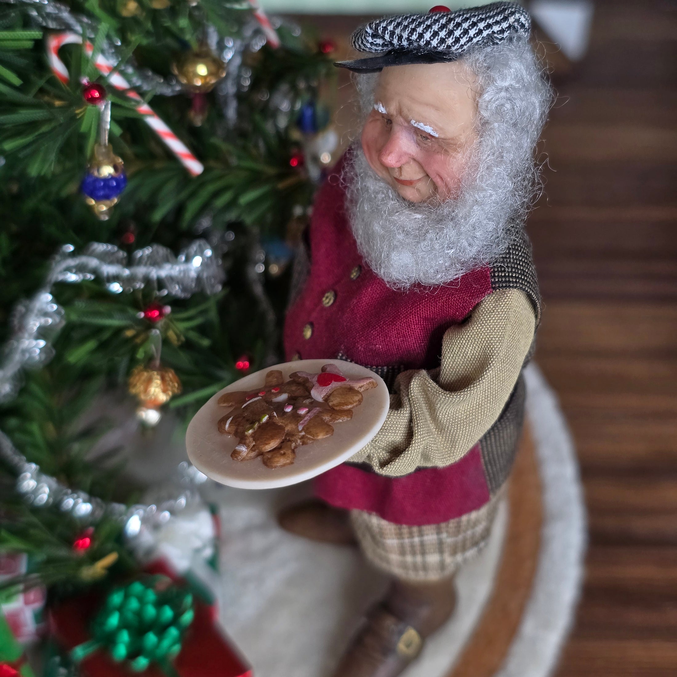 Old Christmas Elf doll with a beard holding cookies in front of a decorated Christmas tree.