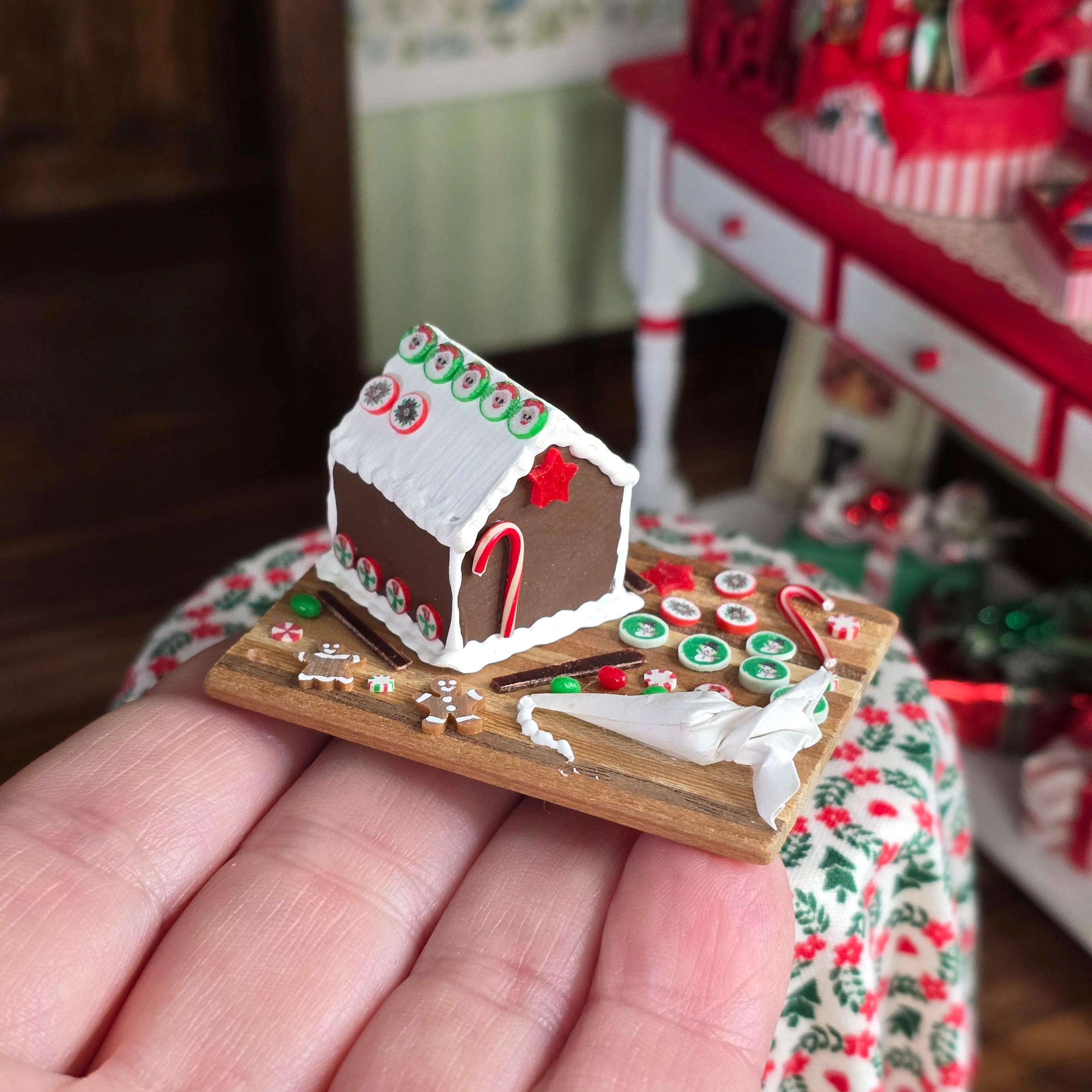 Miniature gingerbread house on a wooden base held in a hand with a blurred festive background.