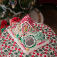 Load image into Gallery viewer, Decorative gingerbread house on a festive tablecloth with a blurred Christmas tree in the background
