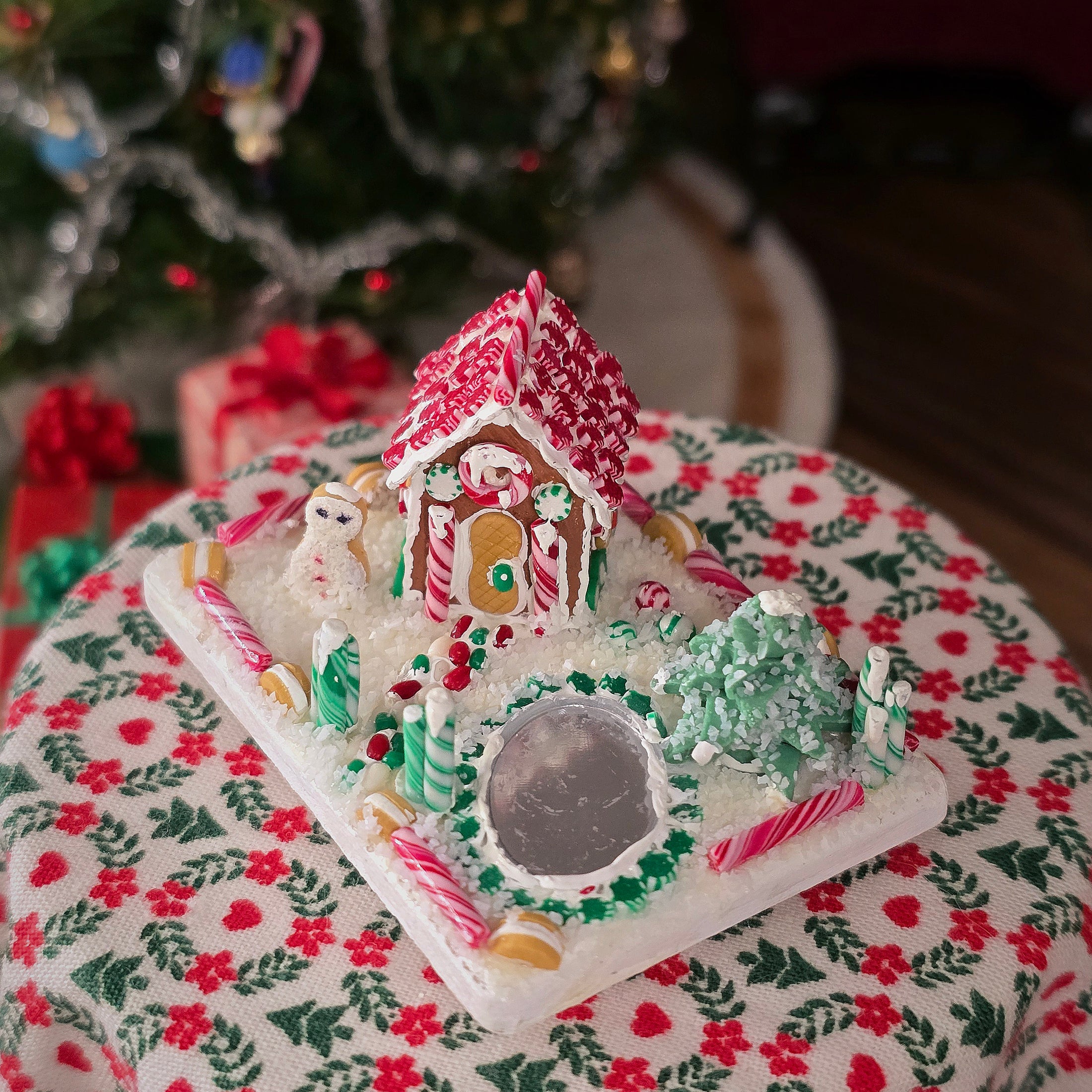 Decorative gingerbread house on a festive tablecloth with a blurred Christmas tree in the background