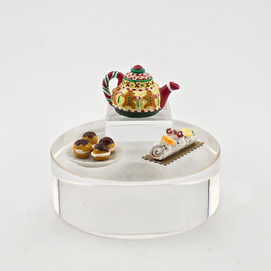 Decorative miniature tea set with a teapot, cake, and pastries on a clear stand against a white background.