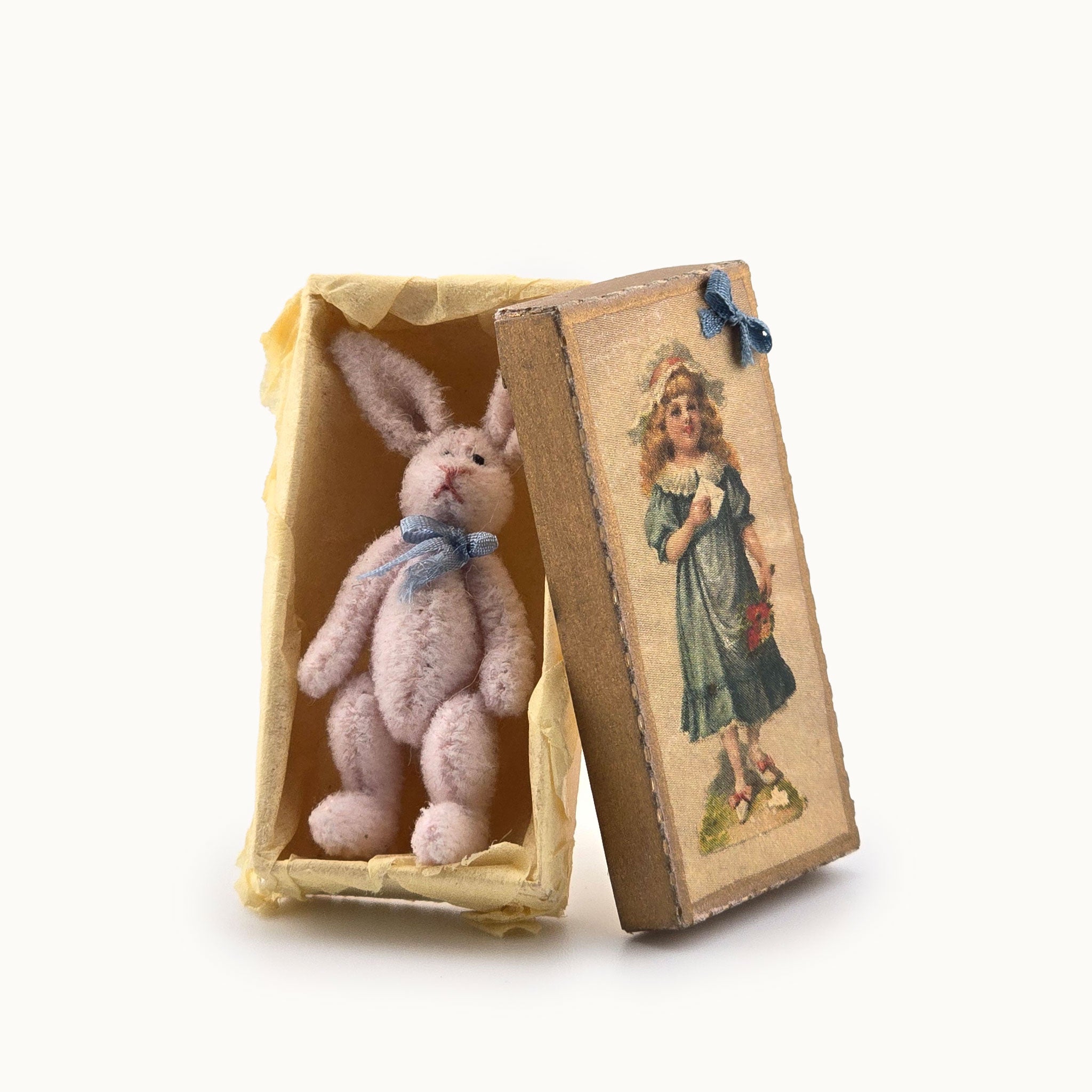 Small pink rabbit toy inside an open vintage-style box with a decorative cover.