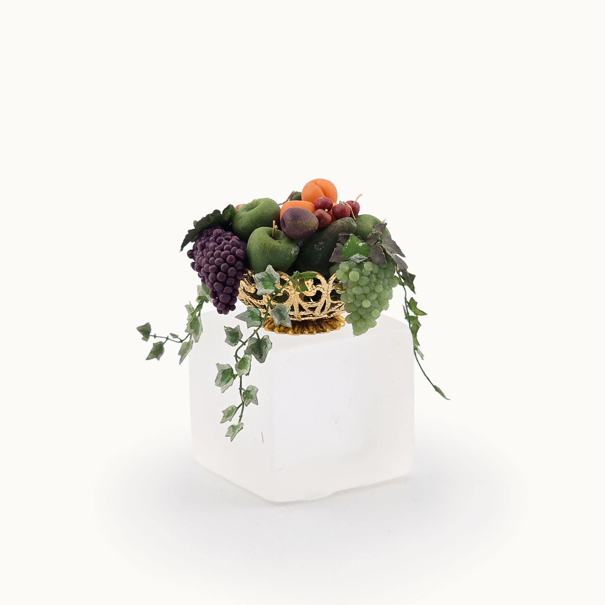 Decorative miniature dollhouse arrangement of fruits and ivy in a brass bowl on a white background